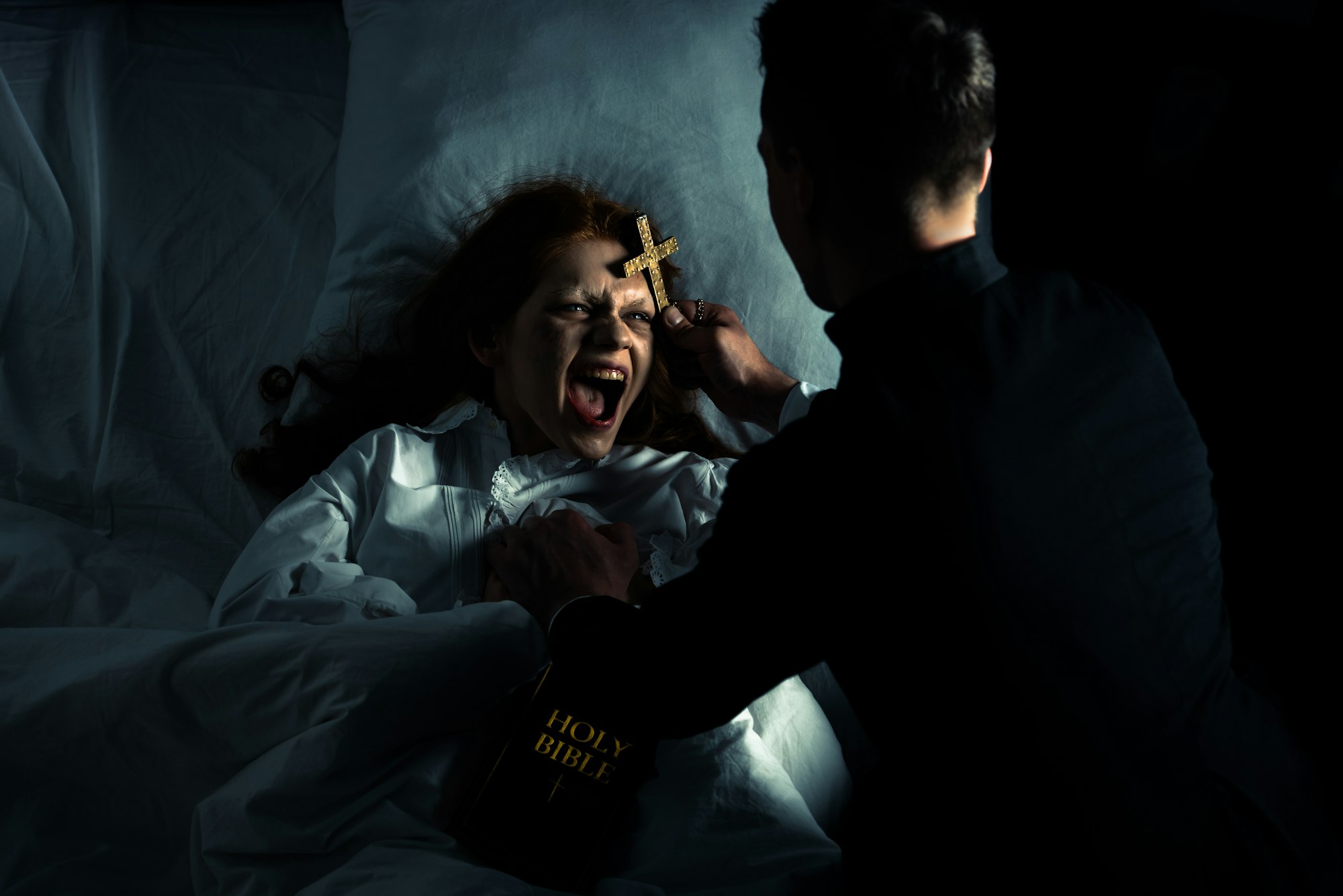 exorcist with bible and cross standing over demoniacal yelling girl in bed